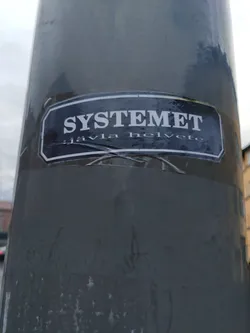 A sticker with the text "SYSTEMET jävla helvete" on a dark gray background. The sticker is affixed to a dark gray pole. The text "SYSTEMET" is in a larger font size than "jävla helvete".