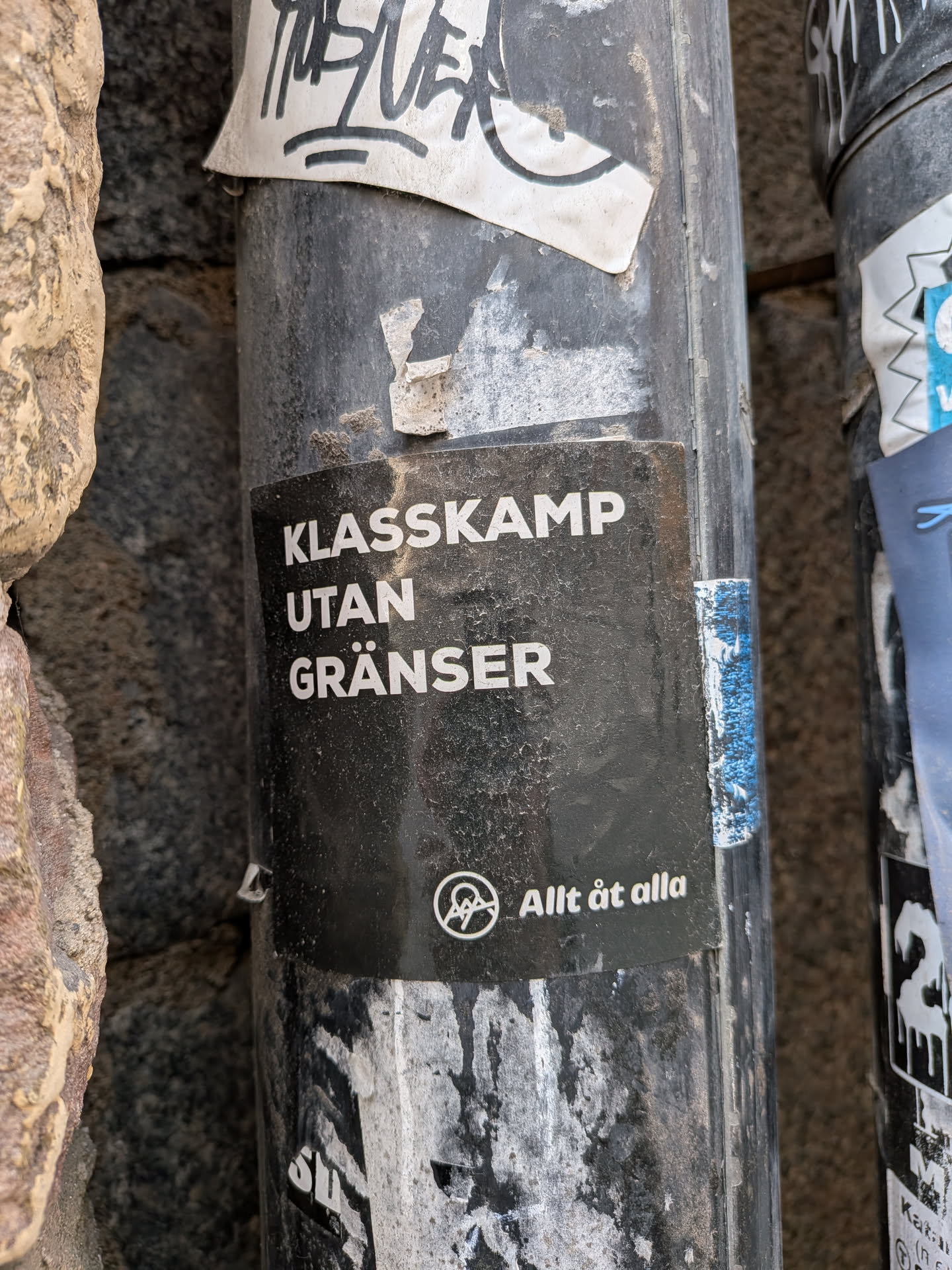 The sticker is rectangular with a solid black background. It features white, sans-serif, uppercase text arranged in three lines: 'KLASSKAMP', 'UTAN', and 'GRÄNSER'. Below this main text, there is a smaller white circular logo, which appears to be a stylized 'AAA' or similar symbol, followed by the text 'Allt åt alla'. The sticker shows signs of wear, with dust and grime on its surface, and some minor peeling at the edges, indicating it has been exposed to the elements on a pole.