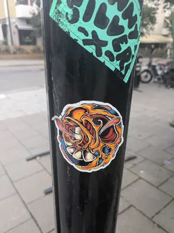 Street sticker The sticker is circular with a distinct white border, applied to a black pole. Its central design features a highly stylized, somewhat abstract face or mask. The dominant colors are fiery oranges and reds, transitioning to yellows, forming flame-like or organic swirling patterns around the central elements. Striking blue accents are present, particularly on the lower left and right, providing a strong contrast. The 'face' itself appears to have large, dark red eye sockets with a white or light-colored, multi-petaled flower-like element at its base, possibly representing a mouth or teeth. The overall impression is dynamic and intense, with bold black outlines defining the various shapes.