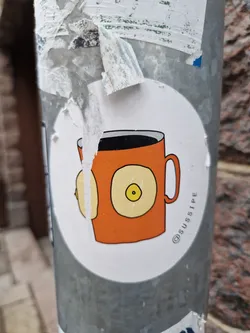 Street sticker A sticker of an orange coffee mug with two breasts on the side. The artist's Instagram handle is @sussipe.
