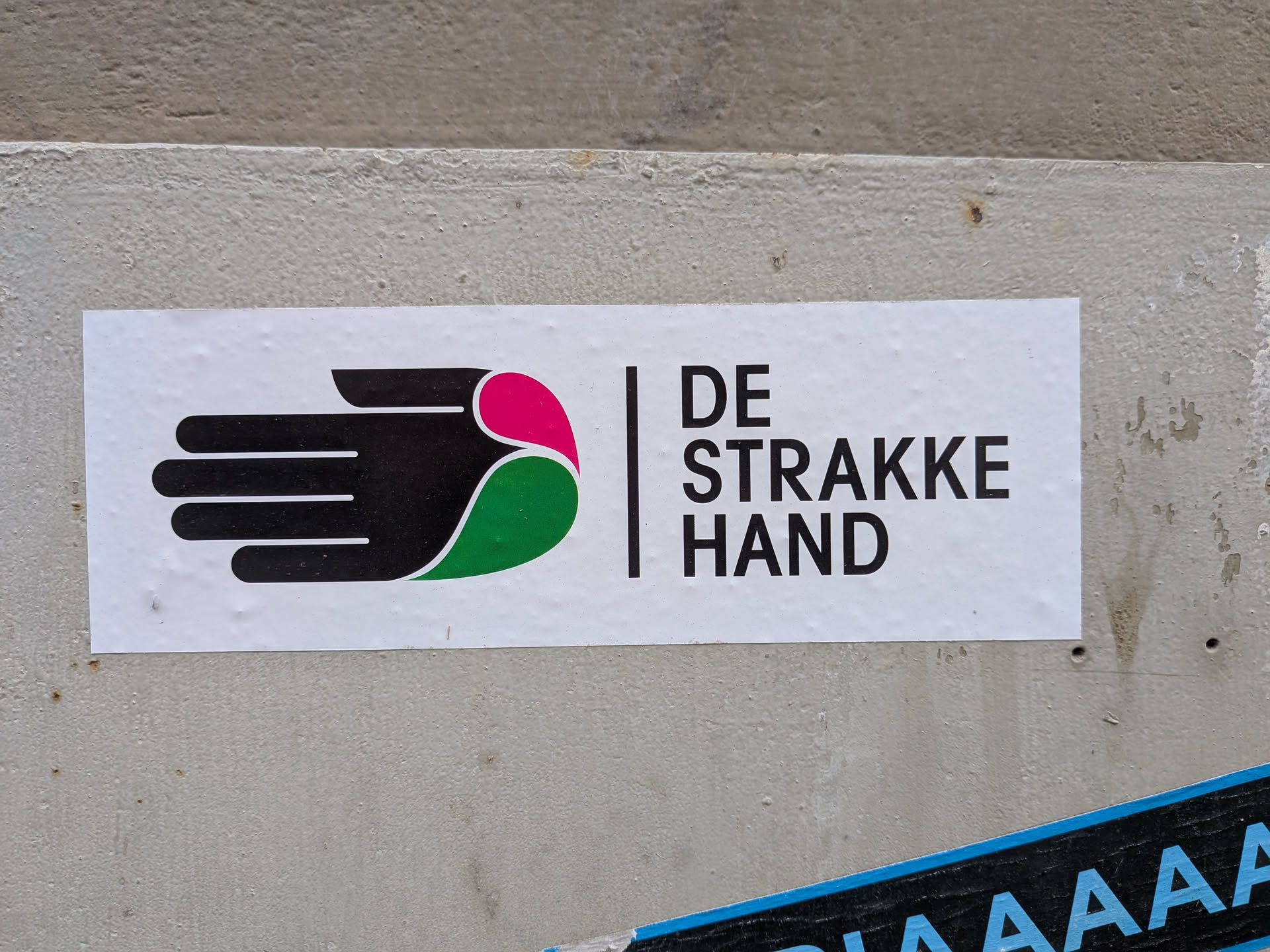 A rectangular white sticker displaying a stylized black hand icon with pink and green accents. To its right, a vertical line separates the icon from the text 'DE STRAKKE HAND' in black, sans-serif font.
