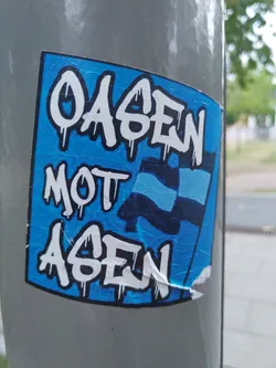 Street sticker A blue sticker with white graffiti-style text. The text says "Oasen mot ägen", which translates from Swedish to "Oases against meadows". There is also a stylized flag in the middle of the sticker. The sticker is slightly peeling at the bottom right corner.