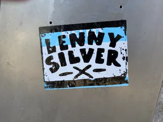 Street sticker A rectangular sticker with a black border, featuring a light blue background. Prominently displayed in the center are the words "LENNY" and "SILVER" in thick, black, hand-drawn block letters. Below "SILVER", there is a stylized black 'X' with two horizontal dashes on either side of it. The edges of the sticker show signs of wear and tear, with some black paint splatter and peeling.