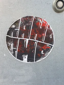 A round sticker with an abstract design. The base is black with red paint splatters, overlaid with a grid of white lines. The style is reminiscent of modern art or street art.