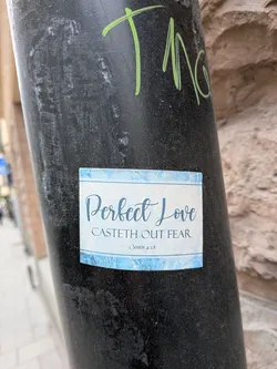Street sticker A rectangular sticker with a light blue, watercolor-like background. The top and bottom edges feature a decorative border with stylized blue floral or leaf patterns. The main text, 'Perfect Love', is written in a large, elegant script font in dark blue. Below it, 'CASTETH OUT FEAR' is in a smaller, all-caps serif font, also in dark blue. At the very bottom, '1 JOHN 4:18' is in a small, simple serif font. The sticker has a clean, spiritual, and calming aesthetic.