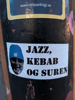 Street sticker The sticker is rectangular with a white background and a slightly wrinkled texture, suggesting it's a physical sticker. On the left side, it features a stylized illustration of a person's face wearing a blue beret. Inside the beret, a small black and white soccer ball design is visible. To the right of the illustration, bold, black, sans-serif text is prominently displayed in three lines: 'JAZZ,', 'KEBAB', and 'OG SUREN'. The text has a stencil-like appearance with small gaps in the letters. The sticker is affixed to a dark, possibly black, cylindrical object that appears weathered and has some reddish marks.