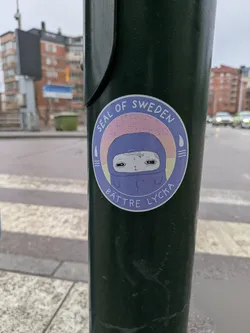 Street sticker A round sticker is affixed to a green pole. The sticker features the text 'SEAL OF SWEDEN' in an arc at the top. Below this, there is an illustration of a character with a pale face and dark eyes, wearing what appears to be a blue hooded garment. The character has a somewhat sad or melancholic expression. The hood is colored in a shade of blue, with a hint of purple around the face. The backdrop consists of a pale orange to pink gradient. There are stylized droplets and wave symbols to the sides of the hooded figure. The text 'BÄTTRE LYCKA' is located at the bottom of the sticker.