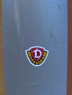 Street sticker A sticker depicting a shield with a stylized letter 'D' inside, above the word 'DRESDEN', affixed to a gray pole.