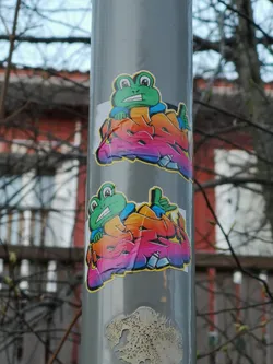 Street sticker The sticker features a cheerful, cartoon-style green frog character with large white eyes and a wide grin showing white teeth. The frog is wearing a blue long-sleeved shirt, and one of its hands is giving a thumbs-up gesture. It appears to be emerging from or integrated with a large, stylized graffiti word or design that transitions in color from orange to pink to purple, outlined in black with white highlights. At the bottom edge of the graffiti design, two small light blue and white spray paint cans are visible. The entire design has a thin yellow outline and a white border, suggesting it's a die-cut sticker.