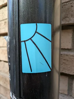 Street sticker The sticker features a simple design of a sun with rays, drawn with black lines on a turquoise background. It is affixed to a black pole. The design suggests a sunny and vibrant theme.