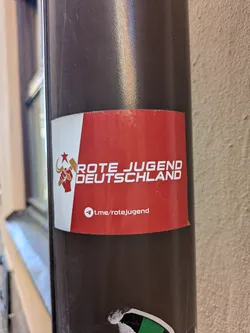 Street sticker A red and white sticker on a pole with the text 'Rote Jugend Deutschland' and a logo featuring a fist holding a hammer and sickle. There is also a link to their Telegram channel.