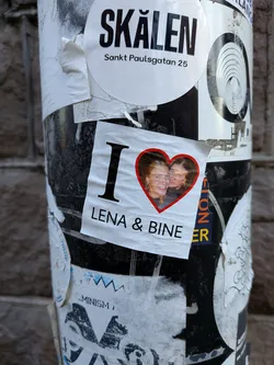 Street sticker A white rectangular sticker featuring a large black 'I' followed by a vibrant red heart. Inside the heart is a color photograph of two smiling individuals, appearing to be a couple. Below the heart, in black capital letters, is the text 'LENA & BINE'. The sticker shows signs of wear, with slight creases and a slightly curled edge, indicating it's a physical sticker affixed to a pole.
