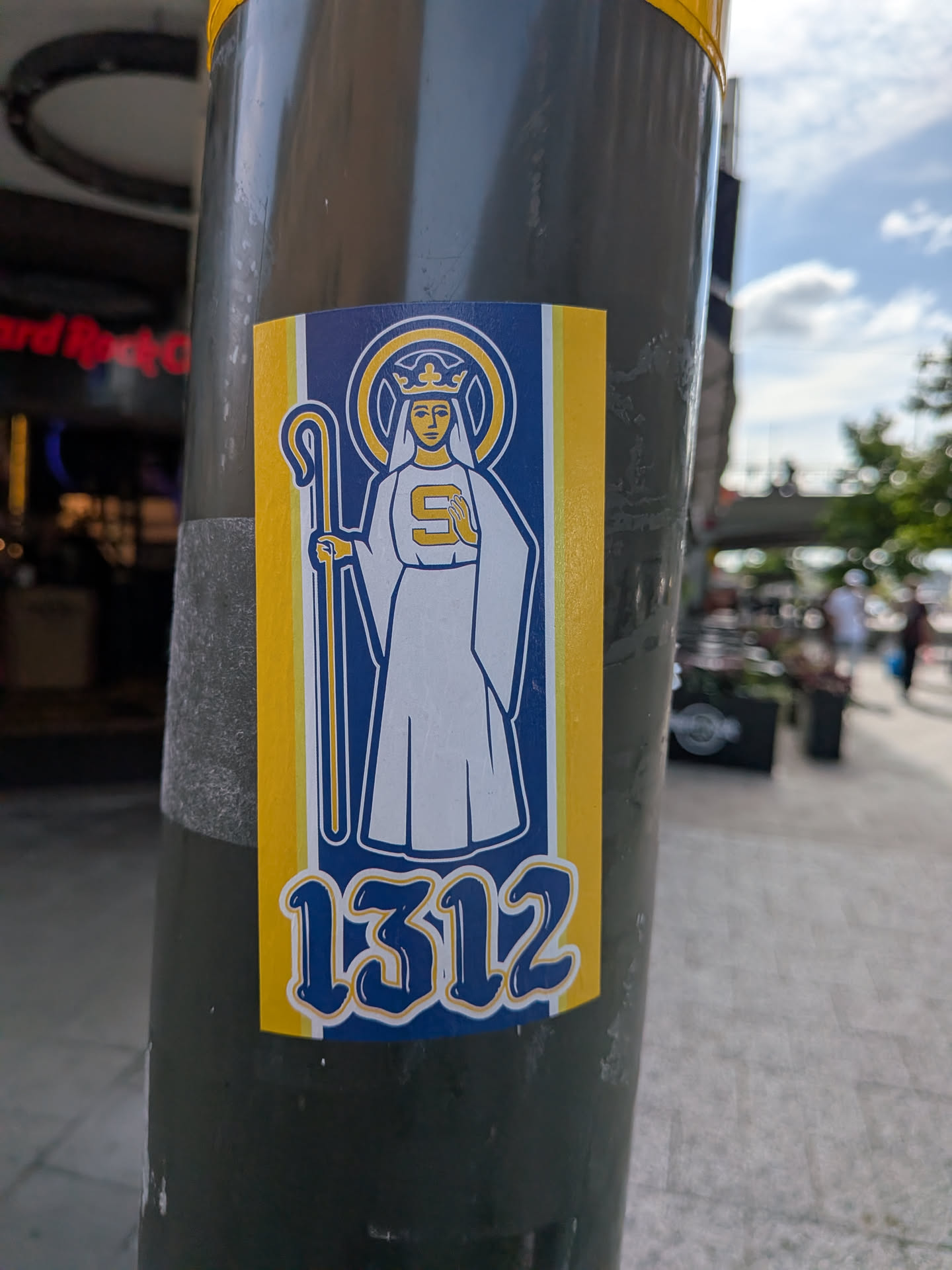 A rectangular sticker depicting a crowned saint with a halo and staff, wearing white robes with an 'S' on the chest. The background has blue and yellow stripes, and the number '1312' is printed at the bottom in a stylized font.