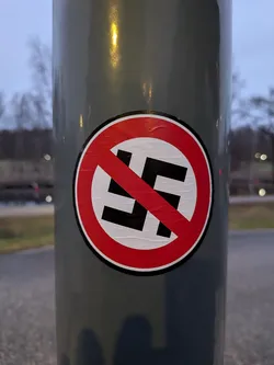 Street sticker The sticker is circular with a red border and a white background. A red diagonal line crosses over a black swastika symbol, indicating prohibition. The sticker appears to be slightly wrinkled, suggesting it is indeed a physical sticker applied to a pole.