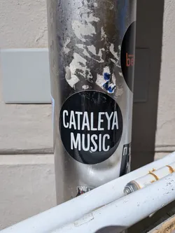 The sticker is a circular, black decal featuring white, sans-serif uppercase text. The text 'CATALEYA' is prominently displayed on the top line, with 'MUSIC' directly below it, both centered within the circle. The sticker has a slight glossy finish and is adhered to a metallic pole, showing minor wear around its edges and reflections from the environment.