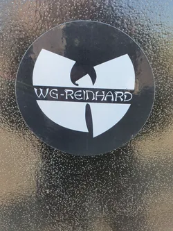 Street sticker A round sticker featuring a stylized Wu-Tang Clan logo with "WG-REINHARD" written underneath. The sticker is black and white and is applied to a textured dark surface. 