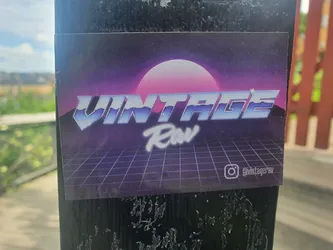 A sticker with a retro 80s design. It features a stylized text saying "VINTAGE Raw" with a grid pattern and a sun in the background. There's an Instagram handle on the sticker.