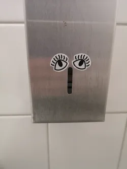 Two stylized eye stickers are placed on a brushed metal dispenser. Each eye is oval-shaped with a thick black outline and a solid black circular pupil. Above each eye, there are several short, curved black lines resembling eyelashes or eyebrows. The background of the eye stickers is white, creating a strong contrast with the black elements. The stickers are positioned symmetrically above a vertical slot in the dispenser, which, in conjunction with the eyes, gives the dispenser an anthropomorphic face.