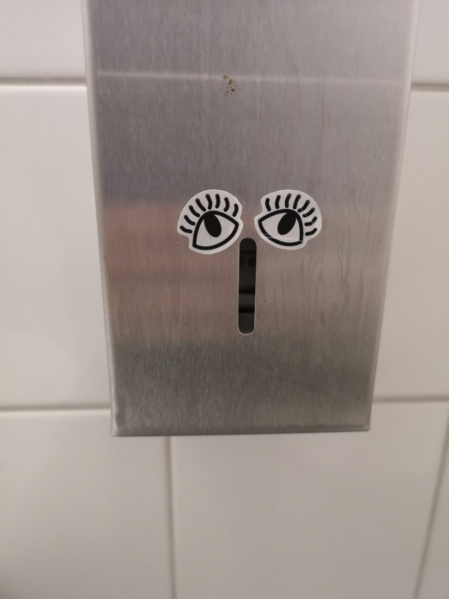Two stylized eye stickers are placed on a brushed metal dispenser. Each eye is oval-shaped with a thick black outline and a solid black circular pupil. Above each eye, there are several short, curved black lines resembling eyelashes or eyebrows. The background of the eye stickers is white, creating a strong contrast with the black elements. The stickers are positioned symmetrically above a vertical slot in the dispenser, which, in conjunction with the eyes, gives the dispenser an anthropomorphic face.