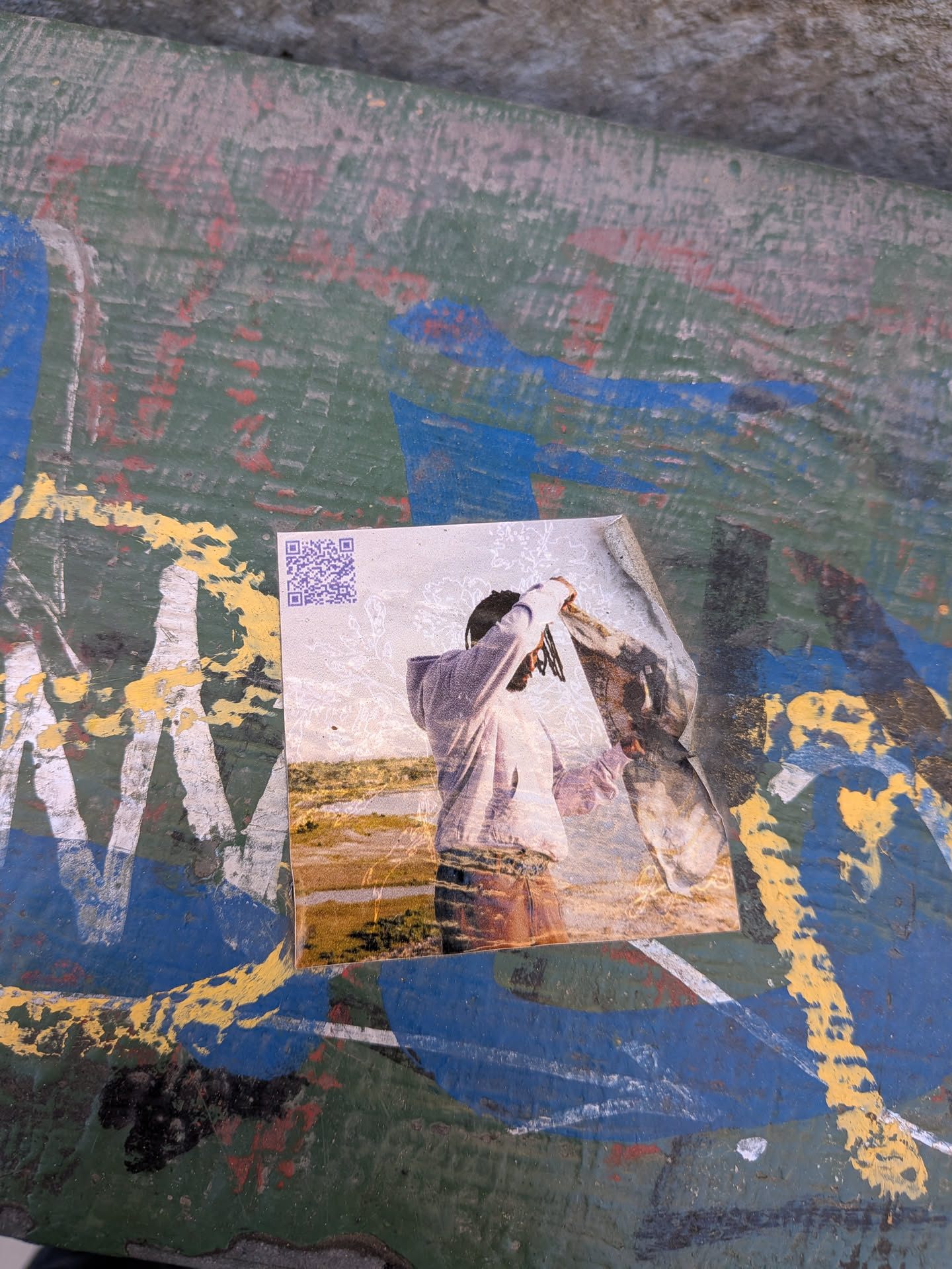A rectangular sticker featuring a photographic image of a person from the waist up, viewed from behind. The person is wearing a light-colored hoodie and appears to be holding up a dark, textured piece of fabric or garment with both hands. Their hair, possibly dreadlocks, is visible under the hood. The background is a natural landscape, likely a marsh or field, with golden-brown grasses and a body of water under a bright sky. A purple QR code is prominently displayed in the top-left corner of the sticker. The sticker itself is slightly worn and peeling at the top right edge, applied to a heavily painted, multi-colored surface with layers of green, blue, yellow, and white paint.