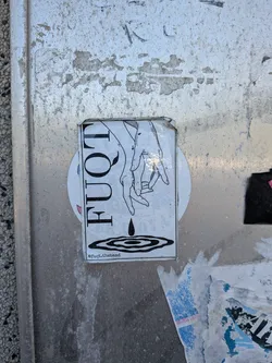 The sticker features a black and white line art illustration. It depicts two hands, one positioned above the other, with fingers gently touching or reaching. From the upper hand, a single tear or water drop falls, creating concentric ripples on a surface below. To the left of the illustration, the stylized text "FUQT" is written vertically in bold, black capital letters. At the very bottom, below the ripples, is the text "@fuqt.theband". The sticker itself is rectangular, with a white background, and is slightly worn with a curled top-right corner, affixed to a textured metallic surface.