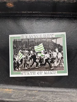 Street sticker A rectangular sticker featuring a black and white photograph framed by a green and white striped border. The photograph depicts a large crowd of people, likely football fans, on what appears to be a stadium pitch or track. Many individuals are running or walking, some holding large green and white striped flags, which are the only elements in color within the photo, standing out against the monochrome background. The background shows stadium stands filled with spectators. Above the photo, in a white banner area, the word "HAMMARBY" is written in bold, outlined capital letters. Below the photo, in a similar white banner area, the phrase "STATE OF MIND" is written in the same style. The overall aesthetic is vintage or retro due to the black and white photo.