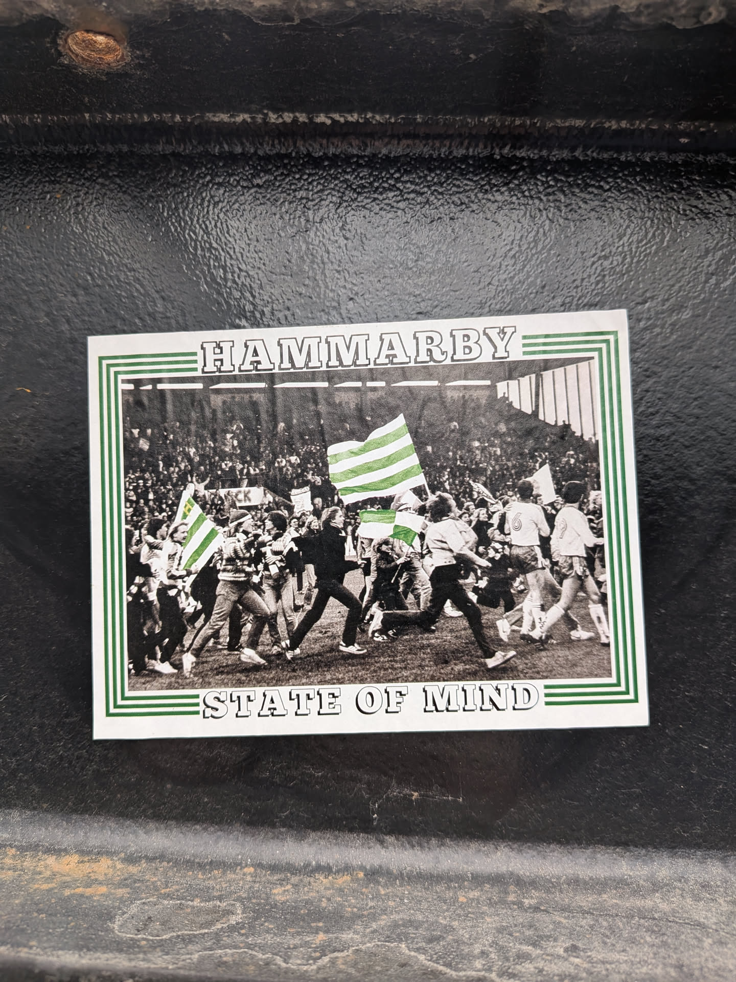A rectangular sticker featuring a black and white photograph framed by a green and white striped border. The photograph depicts a large crowd of people, likely football fans, on what appears to be a stadium pitch or track. Many individuals are running or walking, some holding large green and white striped flags, which are the only elements in color within the photo, standing out against the monochrome background. The background shows stadium stands filled with spectators. Above the photo, in a white banner area, the word "HAMMARBY" is written in bold, outlined capital letters. Below the photo, in a similar white banner area, the phrase "STATE OF MIND" is written in the same style. The overall aesthetic is vintage or retro due to the black and white photo.