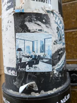 Street sticker The sticker features a rectangular image of a man in a dark suit seated at a large, dark desk in a modern office. He is positioned slightly to the right of the center, looking towards the viewer. Behind him, a large window offers a panoramic view of a city skyline with numerous high-rise buildings. A black desk lamp is visible on the left side of the desk. The image has a cool, desaturated color palette, predominantly blues and greys, giving it a somewhat monochromatic or duotone appearance. The sticker itself is adhered to a pole and shows signs of wear, including some creases and slight fading.