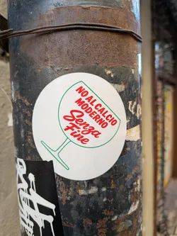 A circular white sticker featuring a stylized green outline of a wine glass. Inside the bowl of the glass, red text is prominently displayed. The upper text, 'NO AL CALCIO MODERNO', is in a bold, sans-serif font, while the lower text, 'Senza Fine', is in a more elegant, cursive script. The sticker is applied to a rusty, dark metal pole.