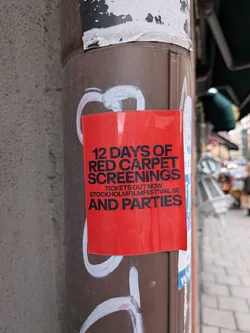 Street sticker A rectangular, glossy red sticker affixed to a brown metal pole. It features bold black sans-serif text advertising '12 DAYS OF RED CARPET SCREENINGS AND PARTIES'. In smaller text in the middle, it reads 'TICKETS OUT NOW' and lists the website 'STOCKHOLMFILMFESTIVAL.SE'. The sticker is pasted over white graffiti markings on the pole.