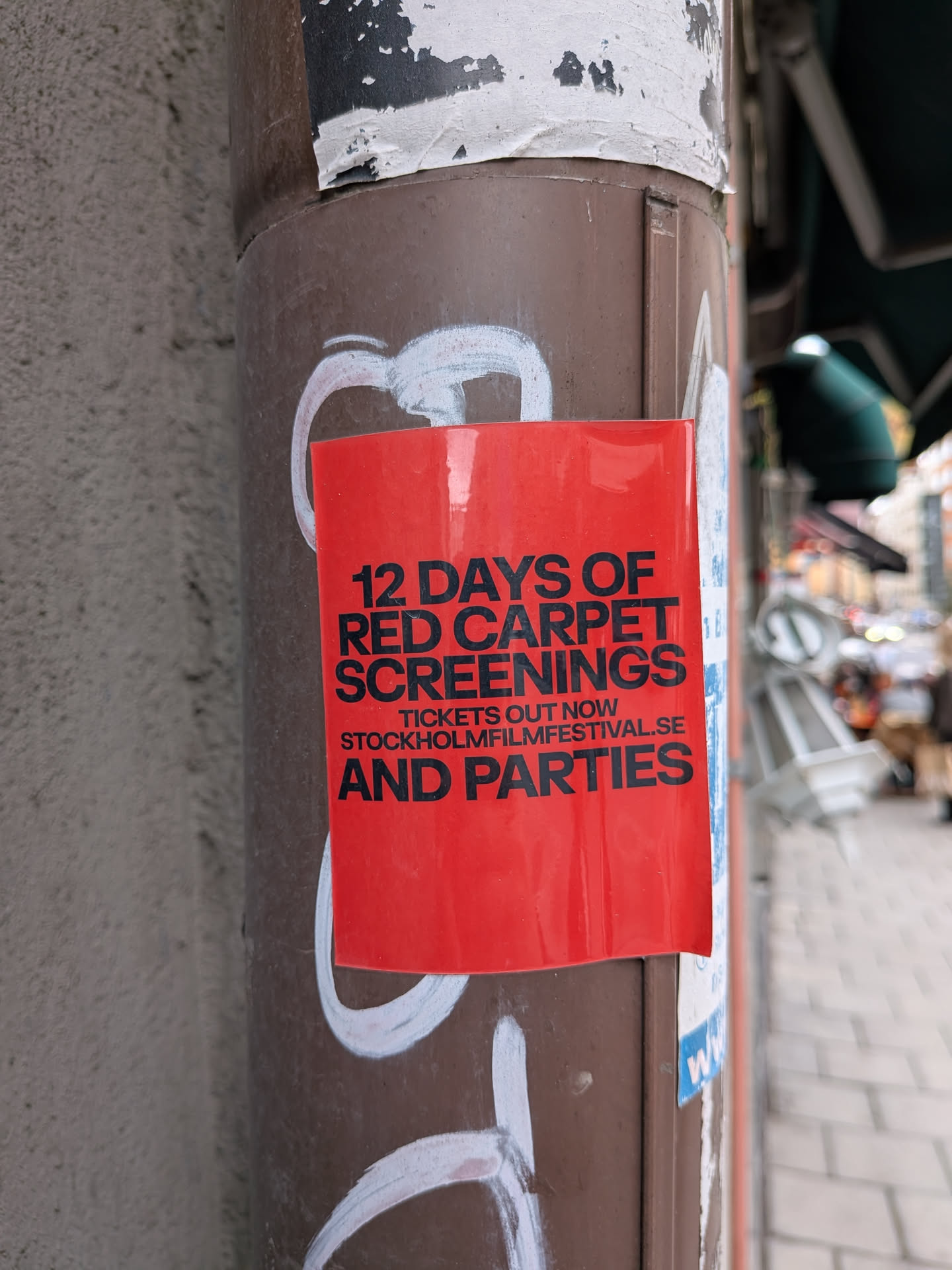 A rectangular, glossy red sticker affixed to a brown metal pole. It features bold black sans-serif text advertising '12 DAYS OF RED CARPET SCREENINGS AND PARTIES'. In smaller text in the middle, it reads 'TICKETS OUT NOW' and lists the website 'STOCKHOLMFILMFESTIVAL.SE'. The sticker is pasted over white graffiti markings on the pole.
