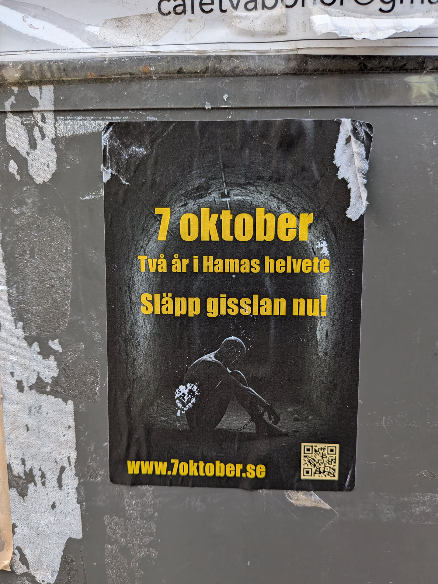 The sticker features a dark, gritty image of what appears to be a tunnel or confined space. In the center, a silhouetted figure is seated, appearing distressed and possibly chained. The text is in bold yellow lettering. The top line reads '7 oktober', followed by 'Två år i Hamas helvete' (Two years in Hamas hell), and 'Släpp gisslan nu!' (Release the hostages now!). A QR code is present in the bottom right corner, and the website 'www.7oktober.se' is displayed below the main text.
