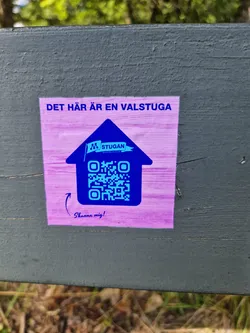 Street sticker Purple and blue house-shaped sticker with a QR code.  The text at the top says "DET HÄR ÄR EN VALSTUGA" (This is a polling station). Inside the house shape is the logo for "M Stugan" and a QR code. At the bottom, it says "Scanna mig!" (Scan me!).
