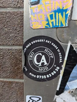 A black and white round sticker adhered to a metal surface. It features the text 'PROBLEM MED DROGER? DET HADE VI OCKSÅ' and displays 'CA MOD' within a circular emblem. Additionally, the sticker contains a phone number: 'RING 0735838630'.