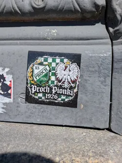 Street sticker A square-shaped sticker featuring a distressed, splattered black background. The central design includes a green and white checkered pattern. On the left, a shield emblem with 'KS PROCH' and '(1926)' in green text is encircled by a golden laurel wreath. On the right, a majestic white Polish eagle with a golden crown, beak, and talons is prominently displayed. Below these elements, the text 'Proch Pionki' is written in a stylized, gothic-inspired font, with '1926' beneath it, all in white. The overall aesthetic combines national symbolism with sports club identity, presented with a rugged, artistic flair.