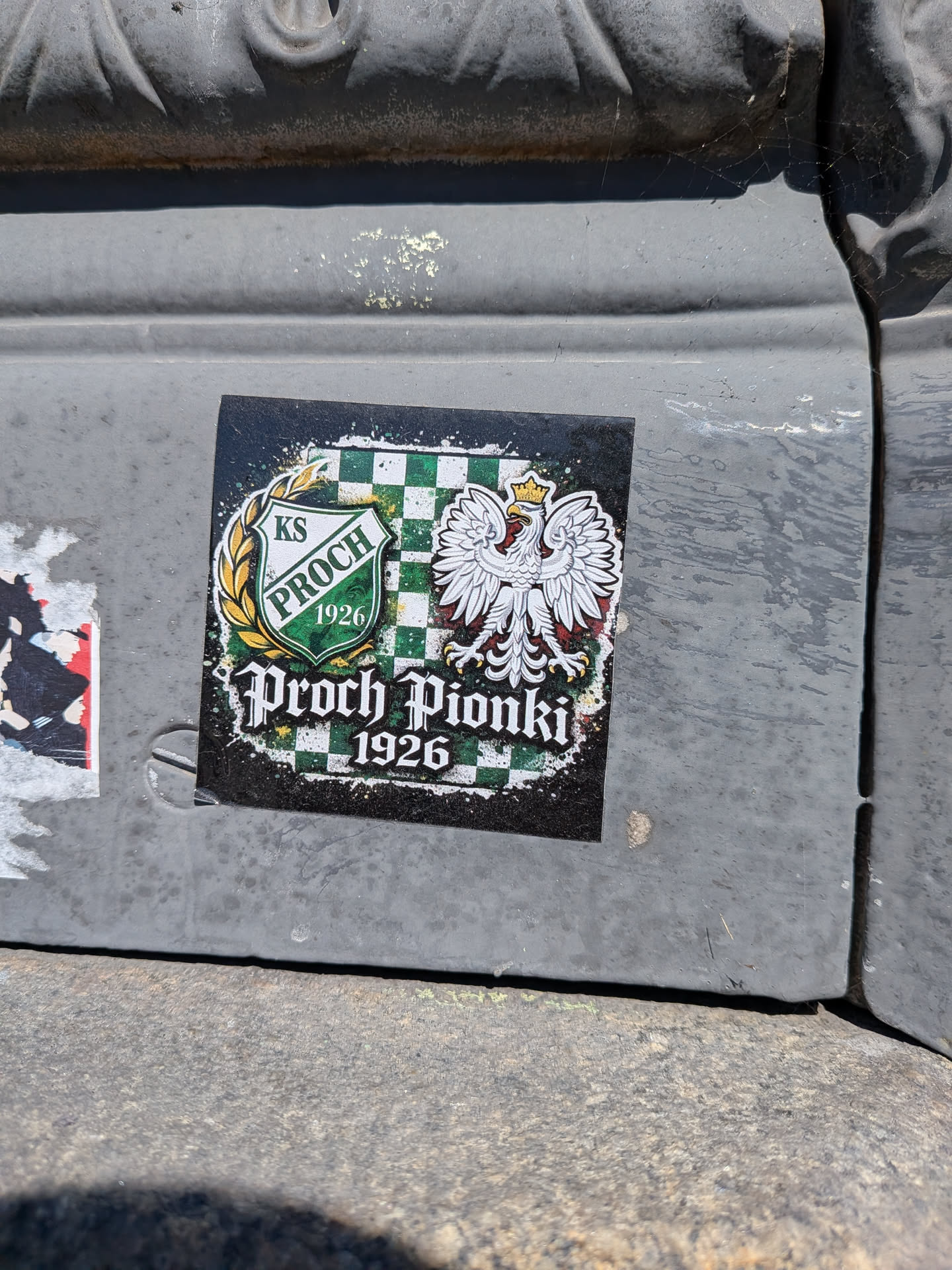 A square-shaped sticker featuring a distressed, splattered black background. The central design includes a green and white checkered pattern. On the left, a shield emblem with 'KS PROCH' and '(1926)' in green text is encircled by a golden laurel wreath. On the right, a majestic white Polish eagle with a golden crown, beak, and talons is prominently displayed. Below these elements, the text 'Proch Pionki' is written in a stylized, gothic-inspired font, with '1926' beneath it, all in white. The overall aesthetic combines national symbolism with sports club identity, presented with a rugged, artistic flair.