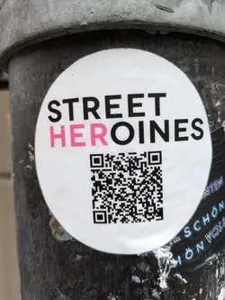 Street sticker Round white sticker with the text STREET HEROINES in black and pink. It contains a QR code in the lower half. The sticker is affixed to a dark gray metallic surface.