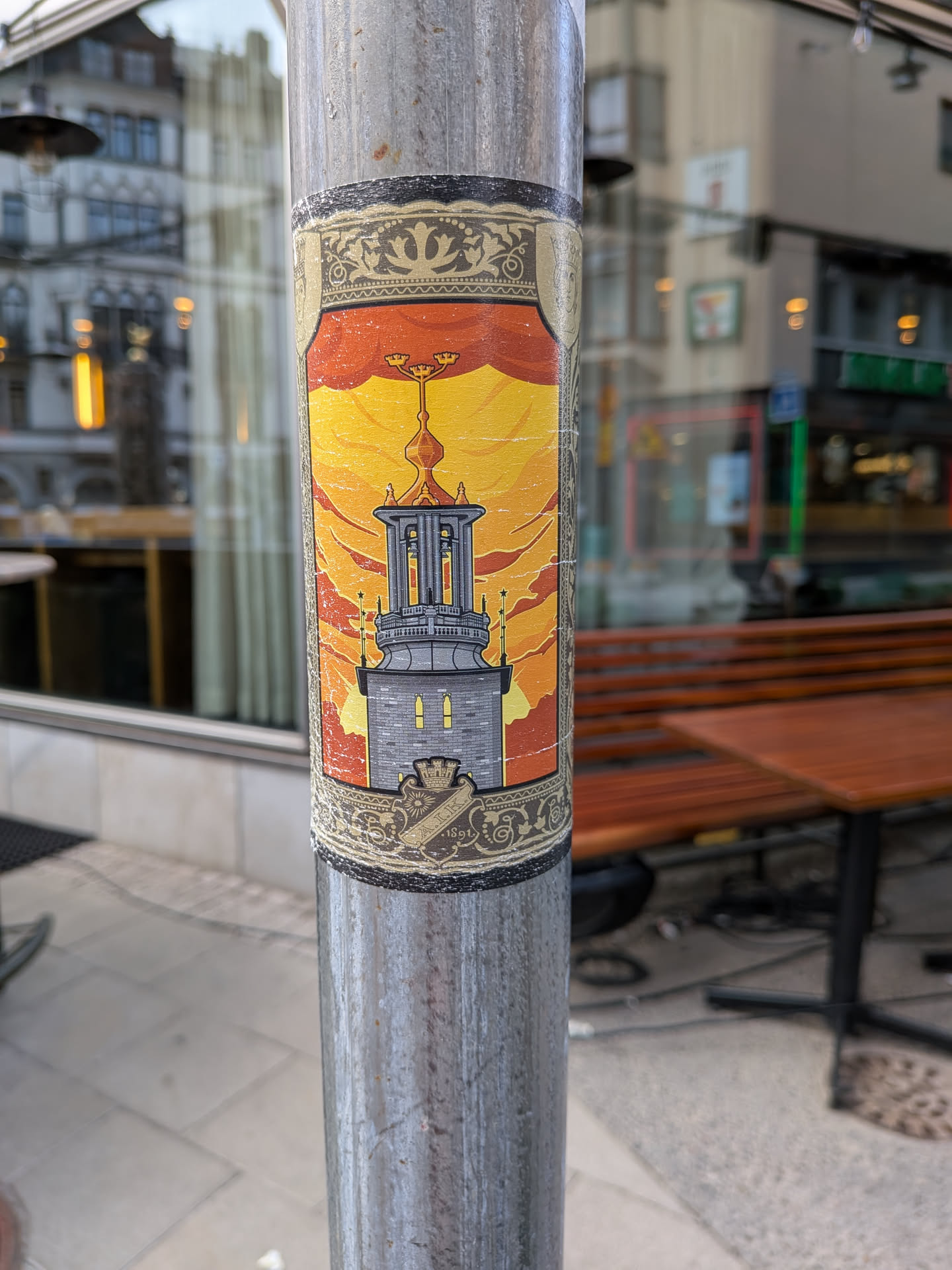 An ornate, vertical sticker on a metal pole. It depicts a stone tower with a golden spire under a yellow-orange sky, framed by gold Art Nouveau borders. The bottom features the AIK crest with '1891'.
