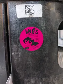 Street sticker A circular, vibrant magenta-pink sticker featuring a black illustration of a cartoon character. The character has a crown or star-like element on its head and is depicted holding a tattoo gun. Its expression is focused or slightly mischievous. Above the character, in bold black capital letters, is the name 'INÊS' with an accent over the 'E', followed by 'LA TATUADORA' in smaller black capital letters. In the bottom right corner, a small social media handle '@INESINK' is visible. The overall style is bold and illustrative, set against a bright pink background.
