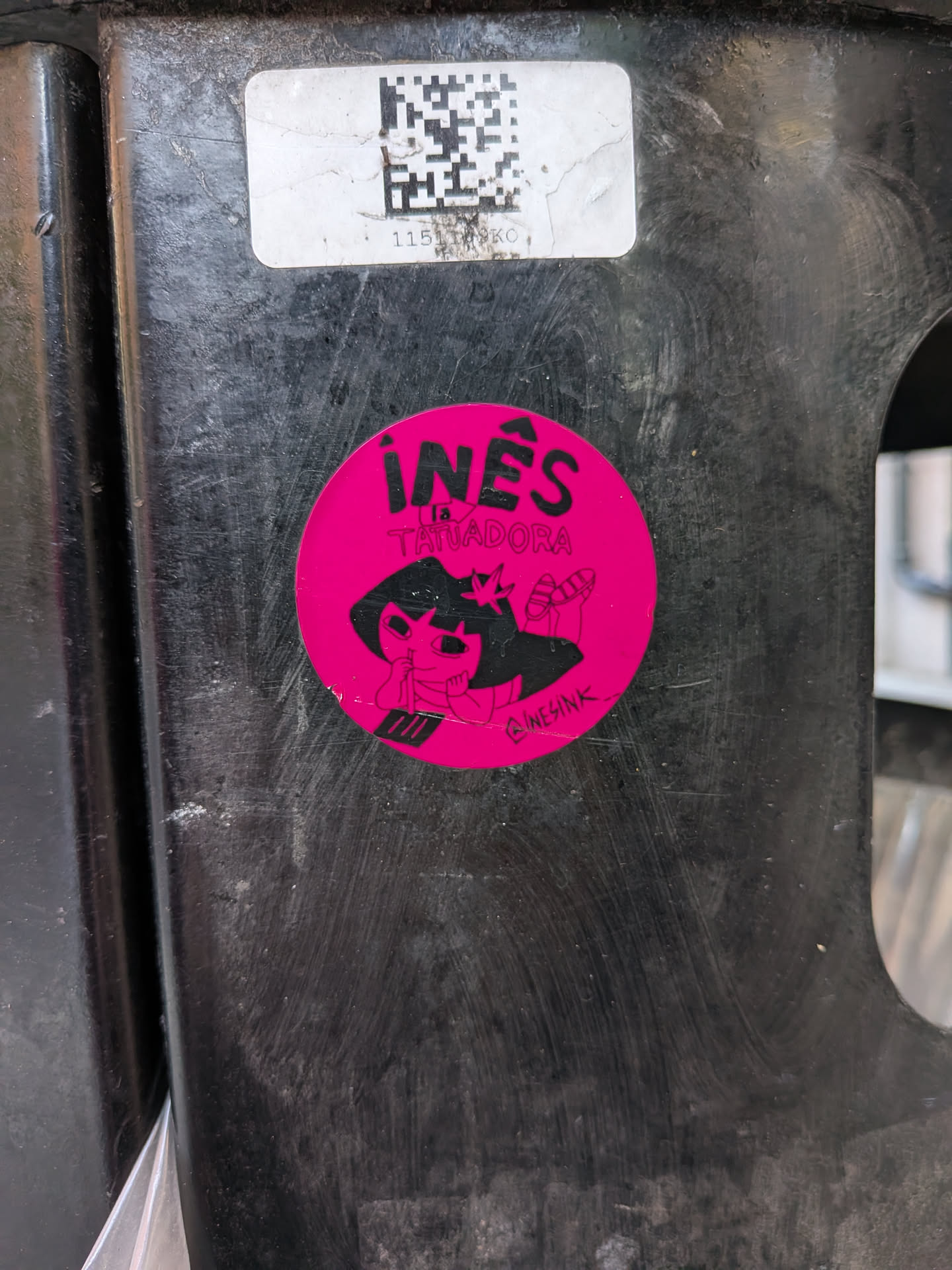 A circular, vibrant magenta-pink sticker featuring a black illustration of a cartoon character. The character has a crown or star-like element on its head and is depicted holding a tattoo gun. Its expression is focused or slightly mischievous. Above the character, in bold black capital letters, is the name 'INÊS' with an accent over the 'E', followed by 'LA TATUADORA' in smaller black capital letters. In the bottom right corner, a small social media handle '@INESINK' is visible. The overall style is bold and illustrative, set against a bright pink background.