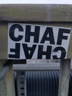 A black and white sticker with the text "CHAFA" written in a bold, sans-serif font. The sticker is affixed to a weathered, light-grey metal surface. The text is vertically inverted. 