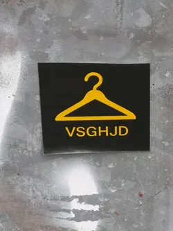 A rectangular sticker with a solid black background. Centered in the upper half is a bright yellow, stylized outline of a clothing hanger. Below the hanger, the capital letters "VSGHJD" are displayed in a bold, sans-serif font, also in bright yellow. The sticker has a clean, high-contrast design against a textured, grey wall.