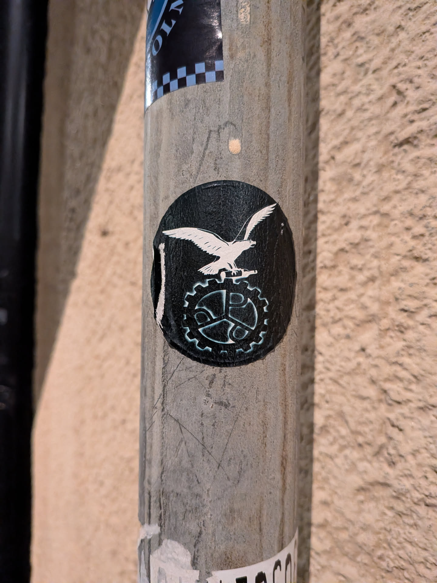 The sticker is circular with a black background. In the center, a white eagle is depicted in flight, with its wings spread. Below the eagle is a stylized gear or cogwheel, also in white, with intricate detailing. The edges of the sticker appear slightly worn and peeling, suggesting it has been affixed for some time.