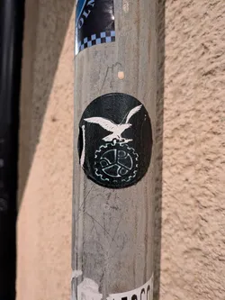 Street sticker The sticker is circular with a black background. In the center, a white eagle is depicted in flight, with its wings spread. Below the eagle is a stylized gear or cogwheel, also in white, with intricate detailing. The edges of the sticker appear slightly worn and peeling, suggesting it has been affixed for some time.