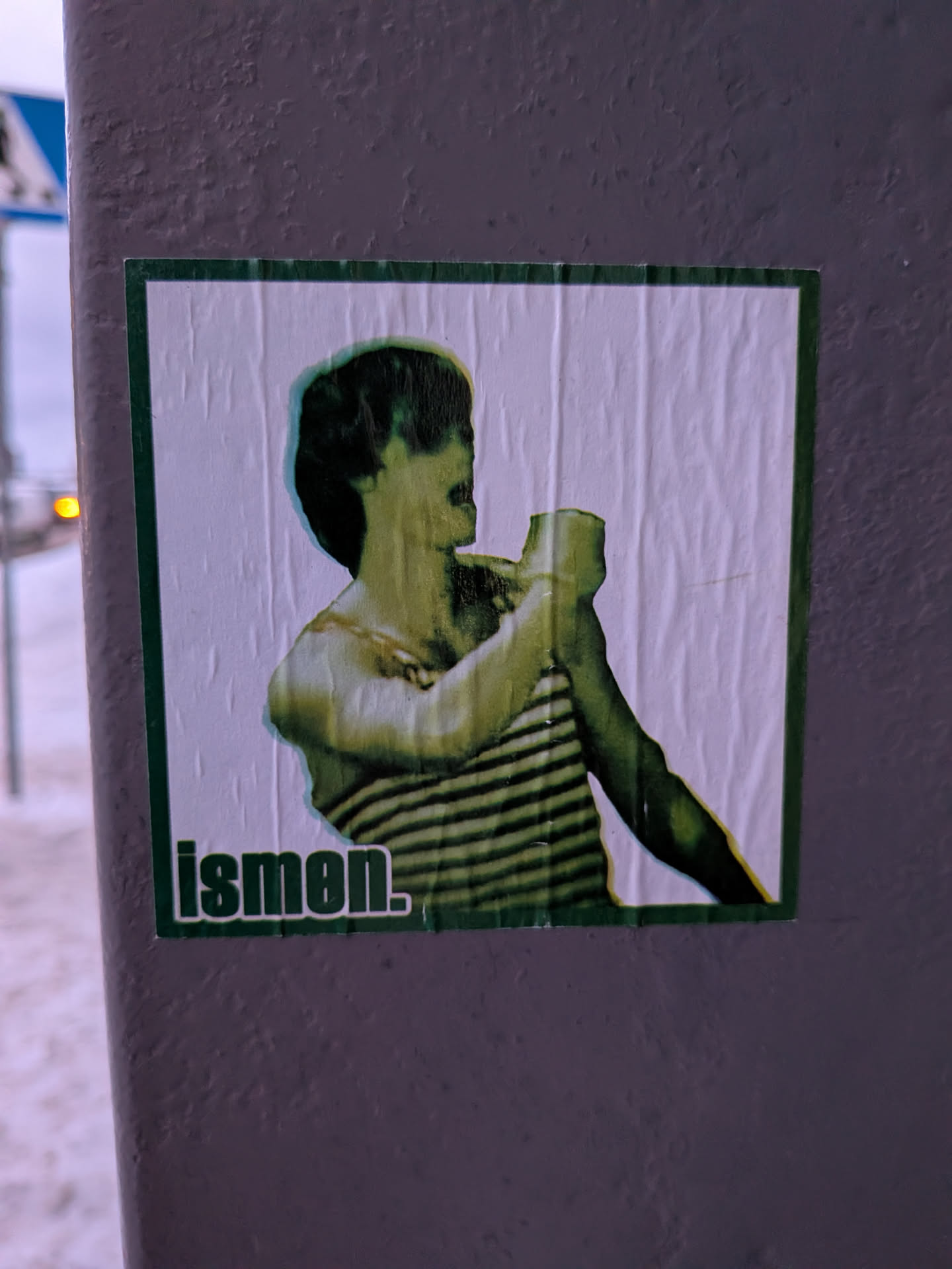 Street sticker A rectangular sticker with a dark green border, featuring a stylized depiction of a person, likely male, from the chest up. The figure is rendered in a distinct greenish-yellow palette with dark green shadows, giving a somewhat distorted or artistic filter effect. They are wearing a horizontally striped shirt and are shown holding an object, possibly a cup or bottle, to their mouth. The background within the sticker is a wrinkled white paper texture. In the bottom left corner, the word "ismen." is printed in a dark green, sans-serif font.