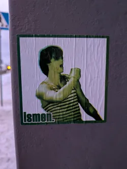 Street sticker A rectangular sticker with a dark green border, featuring a stylized depiction of a person, likely male, from the chest up. The figure is rendered in a distinct greenish-yellow palette with dark green shadows, giving a somewhat distorted or artistic filter effect. They are wearing a horizontally striped shirt and are shown holding an object, possibly a cup or bottle, to their mouth. The background within the sticker is a wrinkled white paper texture. In the bottom left corner, the word "ismen." is printed in a dark green, sans-serif font.