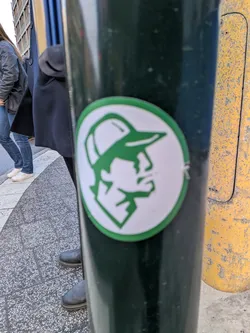 Street sticker The sticker is circular, featuring a thick green outer border enclosing a white background. At the center, a stylized, geometric profile of a person's head, facing right, is rendered in solid green. The figure wears a baseball cap, and facial features like the nose, mouth, and chin are depicted with sharp, angular lines, giving it a minimalist and iconic appearance.