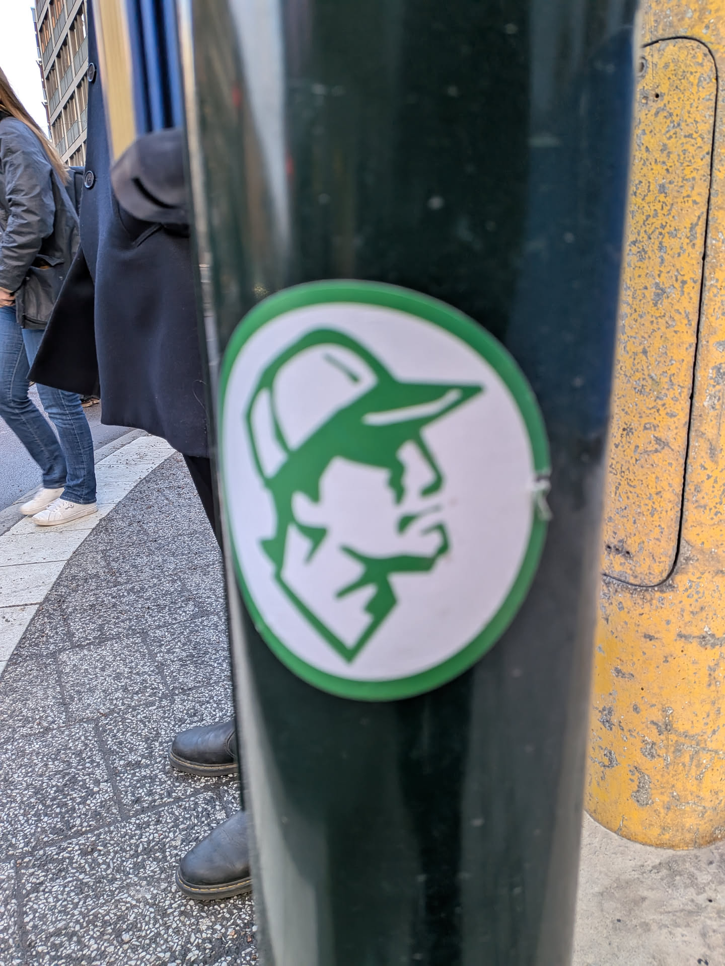 The sticker is circular, featuring a thick green outer border enclosing a white background. At the center, a stylized, geometric profile of a person's head, facing right, is rendered in solid green. The figure wears a baseball cap, and facial features like the nose, mouth, and chin are depicted with sharp, angular lines, giving it a minimalist and iconic appearance.
