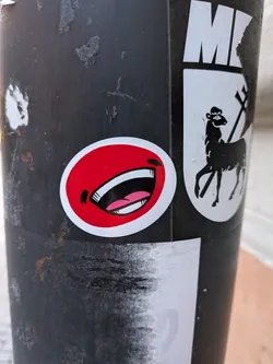 Street sticker The sticker is oval-shaped with a bright red background. It prominently displays a large, open mouth with white teeth and a pink tongue. The mouth is drawn in a cartoonish style with bold black outlines. A small black shape resembling a nose or a shadow is positioned above the mouth. The sticker has a white border.