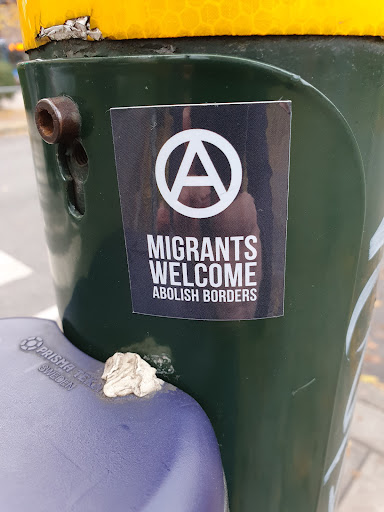 Street sticker A black and white sticker with the anarchy symbol and the text "Migrants Welcome" above "Abolish Borders". It is affixed to a dark green pole.